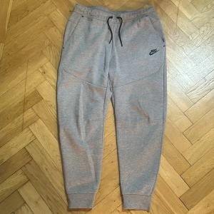 Grå mjukisbyxor från Nike, storlek L - Snygga grå mjukisbyxor från Nike med svart logga på låret. Byxorna har snörning i midjan, muddar vid bensluten och praktiska fickor, varav en med dragkedja bak. Perfekta för chill eller träning, tillverkade i mjukt bomullsmaterial.