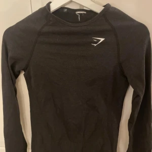 Svart långärmad träningströja Gymshark - Svart långärmad träningströja från Gymshark med vit logga på bröstet. Tight passform och tillverkad i mjukt, stretchigt material som passar perfekt till gymmet eller löprundan. Enkel och stilren design utan extra detaljer.🏃‍♀️‍➡️☀️💗