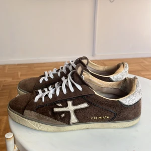 Bruna sneakers från Premiata - Säljer ett par bruna sneakers från Premiata i mocka med vita detaljer och snörning. Skorna har en beige insida och vit, mönstrad hälkappa med sprucken effekt. Klassisk rund tå och platt sula. Perfekta för dig som gillar stilrena sneakers. Köpte dem med gråa skosnören, men bytte ut dem för vita. Men självklart får ni med original skosnören också. För fler bilder, frågor eller prisförslag är det bara att skriva! 😁Priset är inte fast.