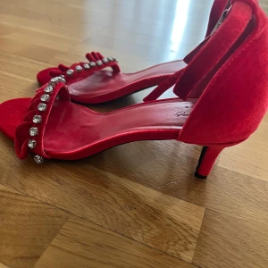 Röda sammetsklackar med strass - Röda pumps i sammet med låg klack och öppen tå. Skorna har en rem runt ankeln och en volangprydd rem framtill dekorerad med glittrande strass. Perfekta för dig som vill sticka ut med färg och detaljer.