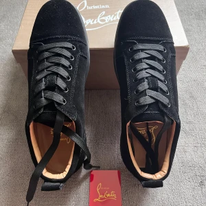 Christian Louboutin svarta skor mocka - Exklusiva svarta skor från Christian Louboutin i mjuk mocka med klassiska röda sulor och svarta snören. Snygg och stilren design med rund tå och diskreta metalldetaljer på sidan. Perfekta för dig som vill sticka ut med lyxig streetstyle. Allt i bilden medföljer