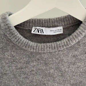 Grå stickad kortärmad tröja från Zara - Supermjuk grå stickad tröja från Zara med korta ärmar och rund hals. Tröjan har ribbade kanter vid hals, ärmslut och nederkant. Perfekt att styla med jeans eller kjol för en chill och trendig look.