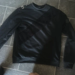 Svart sweatshirt från Le Vichy XS - Svart sweatshirt från Le Vichy i storlek XS. Tröjan har en unik diagonal sammetsdetalj framtill, ribbade muddar och coola spännen på axlarna. På vänster ärm sitter en svart patch med Le Vichy-logga. Perfekt för dig som gillar stilrena och moderna plagg.
