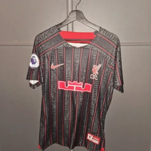 Liverpool FC x Lebron - Liverpool FC fotbollströja i svart med röda och grå vertikala ränder, Premier League-märke på ärmen och Nike-logga på bröstet. Tröjan har klubbens emblem och röd detalj vid halsen. Tillverkad i lätt och ventilerande syntetmaterial.
