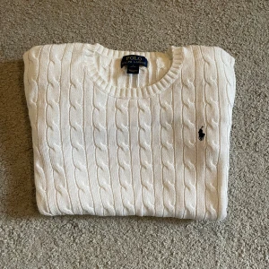 Vit kabelstickad tröja Polo Ralph Lauren - Vit kabelstickad tröja från Polo Ralph Lauren med klassisk rund halsringning och broderad marinblå logga på bröstet. Tröjan har långa ärmar och ribbade muddar vid ärmslut och nederkant. Perfekt för lager-på-lager och snygg till jeans.