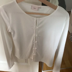 Vit ribbad topp med rosetter från Nelly - Supersöt vit ribbad topp från Nelly med långa ärmar och flera små rosetter framtill. Stretchigt material som sitter snyggt och känns bekvämt. Perfekt att styla med jeans eller kjol för en clean look. Helt ny