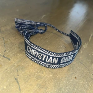 Christian Dior armband - Snyggt vävt armband från Christian Dior i marinblått och vitt med texten 'CHRISTIAN DIOR' och 'DIOR J'ADIOR'. Armbandet har justerbar knytning med tofsar i ändarna och cool kontrastkant. Perfekt statement-piece för dig som gillar lyxiga accessoarer.