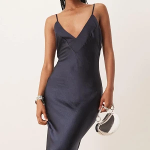 Slutsåld mörkblå långklänning i satin - Slutsåld elegant mörkblå långklänning i glansigt satinmaterial med smala axelband, v-ringning och en snygg rosett i midjan från märket SNDYS. Slutsåld på hemsidan! Perfekt för dig som vill sticka ut med en stilren look. Endast använd en gång. Ordinarie pris: 909kr