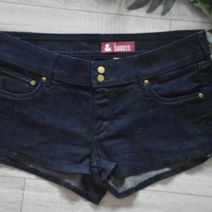 Mörkblå jeansshorts med guldknappar - Supersnygga mörkblå jeansshorts med låg midja och guldiga knappar framtill. Klassisk femficksmodell med kontrastsömmar och korta ben. Perfekta för varma dagar när du vill ha en cool och avslappnad look.