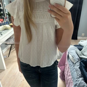 Vit blus med spets från Vero Moda - Supersöt vit blus från Vero Moda med volangärmar och broderade spetsdetaljer. Blusen har små prickar i tyget, rund hals med rysch och knapp i nacken. Luftig och lätt i bomull, perfekt för vår och sommar.