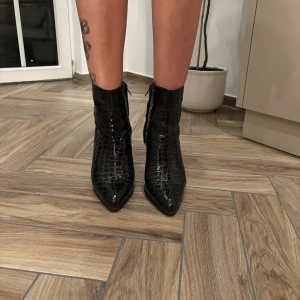 Svarta boots med krokopräglat mönster - Snygga svarta boots från Cobbler i storlek 40 med krokopräglat mönster. Skorna har spetsig tå, hög klack och dragkedja på sidan. Tillverkade i glansigt syntetmaterial som ger en lyxig känsla. Perfekta för dig som vill sticka ut med en trendig look. Klacken är 9cm ungefär. Säljer för att klacken är för hög för min smak. 