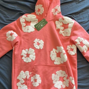 Rosa hoodie från Denim Tears med blommor - Säljer en rosa hoodie från Denim Tears med stora vita blommiga mönster över hela tröjan. Tröjan har huva, känguruficka och är gjord i 100% bomull. Perfekt statement-plagg för dig som vill sticka ut med cool design och färg. Priset kan diskuteras!😁