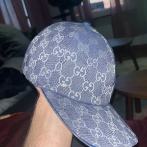 Blå GG-keps från Gucci i jeans - Säljer en blå keps från Gucci i jeansmaterial med ikoniskt GG-mönster över hela kepsen. Klassisk böjd skärm och snygg passform. Perfekt för dig som vill sticka ut med en lyxig accessoar från ett av världens största modehus.