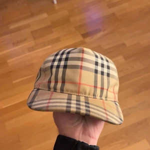 Burberry rutig keps i beige och svart - Snygg keps från Burberry med klassiskt rutigt mönster i beige, svart, vitt och röda linjer. Justerbar snapback baktill och tydlig Burberry London England-patch. Perfekt accessoar för att lyfta din outfit med lyxig streetkänsla.