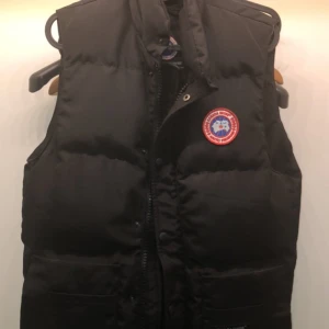 Canada Goose väst  - Säljer en svart dunväst från Canada Goose med ett emblem på bröstet. Västen har en dragkedja och knappar framtill för extra skydd mot kyla. Perfekt för kyliga dagar.