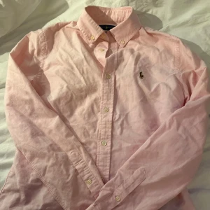 Rosa skjorta från Ralph Lauren - Säljer en stilren rosa skjorta från Ralph Lauren i slim fit. Skjortan har långa ärmar och knappar framtill samt en broderad logotyp på bröstet. Perfekt för en elegant look. Det är en liten fläck men som är rätt så diskret men annars mycket bra.