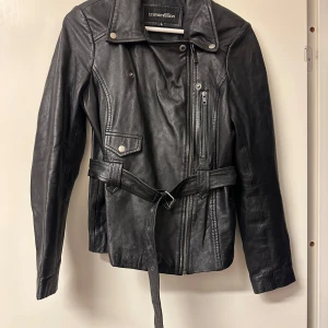 Svart skinnjacka från Transmission - Snygg svart skinnjacka från Transmission med dragkedja och bälte i midjan. Jackan har långa ärmar och flera fickor med dragkedjor. Perfekt för en cool och stilren look.