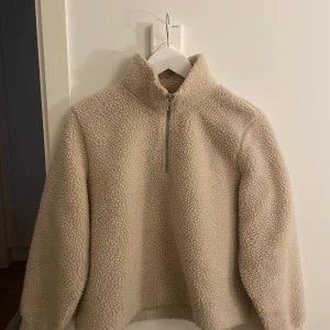 Beige teddyjacka med dragkedja - Mysig beige teddyjacka med hög krage och dragkedja framtill. Perfekt för kyliga dagar med sin fluffiga och varma design. Jackan har långa ärmar och en avslappnad passform.