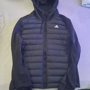 Svart pufferjacka från Adidas - Säljer en svart pufferjacka från Adidas med huva och dragkedja. Jackan har långa ärmar och är perfekt för kyligare dagar. Den har en stilren design med Adidas-loggan på bröstet.