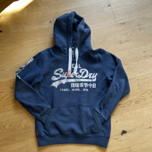 Blå hoodie från Superdry - Säljer en blå hoodie från Superdry med text på bröstet och ärmen. Den har en klassisk känguruficka och justerbar huva med vita snören. Perfekt för en avslappnad stil.