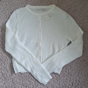 Vit ribbad kofta från 157 - Säljer en stilren vit ribbad kofta från 157. Koftan har långa ärmar och knappar framtill, den är mjuk och stretchig. Perfekt för lager-på-lager och passar till många olika outfits. 