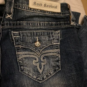 Blå jeans med broderade detaljer - Snygga blå jeans med slitningar och broderade detaljer på bakfickorna. Från rock revival. Säljer för att de inte passar mig. Pris kan diskuteras, Skriv om ni har frågor!💕