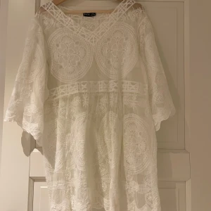 Vit spets coverup  - Säljer en vacker vit cover up med ett detaljerat broderat mönster. Klänningen har en v-ringad hals och trekvartsärmar, perfekt för sommarens stränder. Den är luftig och elegant, vilket gör den till ett måste i garderoben. Passar perfekt för en dag på stranden eller en somrig strandfest.