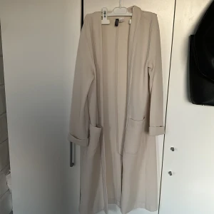 Beige lång kofta från H&M - Säljer en stilren beige lång kofta från H&M. Den har långa ärmar och två praktiska fickor framtill. Perfekt för en elegant look.