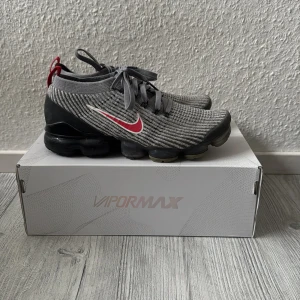 Nike Vapormax Flyknit 3 i grått och rött - Snygga Nike Vapormax Flyknit 3 sneakers i grått med röda detaljer. Skorna har en stickad ovandel och en unik sula med synliga luftkuddar för extra komfort. Perfekta för både stil och funktion. Bra kondition, nypris runt 2000