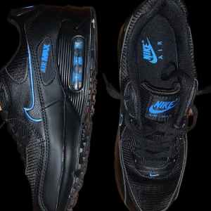 Nike Air Max 90 - Snygga Nike Air Max sneakers i svart med blå detaljer. Skorna har en ikonisk luftenhet i sulan för extra komfort och stil. Perfekta för både vardag och träning.