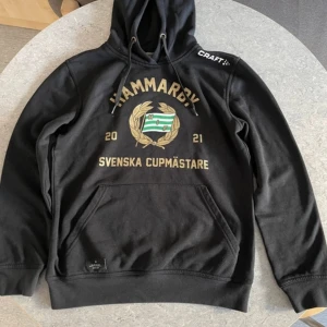 Svart hoodie från Craft med Hammarby-tryck - Säljer en svart hoodie från Craft med Hammarby-tryck och texten 'Svenska Cupmästare 2021'. Tröjan har en stor ficka framtill och justerbar huva med dragsko. Perfekt för alla Hammarby-fans! 🖤