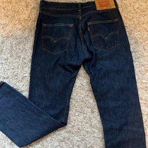 Levis 501 jeans i mörkblå denim - Klassiska Levis 501 jeans i mörkblå denim. Dessa jeans har en rak passform och är tillverkade i slitstarkt denimtyg. De har fem fickor och en knappgylf. Perfekta för en tidlös stil.