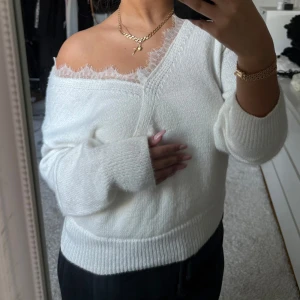 Vit stickad offshoulder tröja - Säljer en stilren vit stickad tröja med offshoulder-design och spetsdetaljer vid halsringningen. Tröjan har långa ärmar och ribbade muddar vid ärmslut och nederkant.