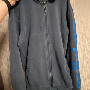 Blå hoodie från Adidas - Säljer en blå hoodie från Adidas med dragkedja och tryck på ärmen. Perfekt för en avslappnad stil. Den har en bekväm passform och en praktisk huva. Saknar snöre runt luvan men annars bra skick. 