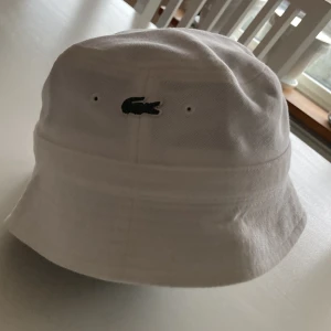Vit bucket hat från Lacoste - Snygg vit bucket hat från Lacoste med det klassiska krokodilemblem broderat framtill. Hatten är tillverkad i bomull och har en stilren design som passar perfekt för soliga dagar.