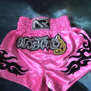 Rosa boxningsshorts från Another Boxer - Snygga rosa boxningsshorts från Another Boxer med svart och guld broderad text och tribal-mönster. De har en elastisk midja för en bekväm passform. Perfekta för träning eller tävling!