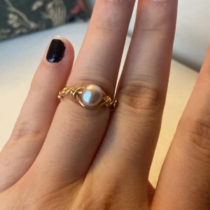 Guldfärgad ring med pärla - Elegant guldfärgad ring med en vacker pärla i centrum. Ringen har en unik design med tvinnade detaljer som ger den en extra touch av stil. Perfekt för att lägga till en sofistikerad känsla till din look.