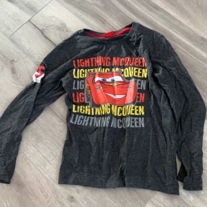 Grå långärmad tröja med Lightning McQueen - Säljer en grå långärmad tröja med tryck av Lightning McQueen från Disney Pixar Cars. Tröjan har färgglada bokstäver i rött, gult och grått samt en röd kant vid halsen. Perfekt för små bilentusiaster. * Barnsize