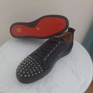 Christian Louboutin  Svarta sneakers - Snygga svarta sneakers från Christian Louboutin med nitar på tån och en ikonisk röd sula. Skorna har snörning och är tillverkade i ett glansigt material som ger en lyxig känsla.