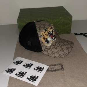Gucci keps med tigerbroderi - Snygg Gucci keps med ikoniskt GG-mönster och broderade tigrar framtill. Kepsen har en justerbar rem baktill med klassiska Gucci-färger. Perfekt för att ge din outfit en lyxig touch. Finns i S & M & L