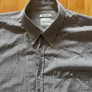 Rutig kortärmad skjorta från Part Two - Säljer en klassisk rutig skjorta från Part Two i storlek XL. Skjortan har korta ärmar och en bröstficka med knapp. Perfekt för en avslappnad stil. Den är i en neutral färgskala med små rutor i svart och vitt.