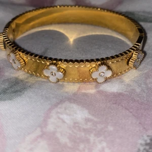 Guldarmband med vita blommor - Elegant guldarmband dekorerat med vita blommor runtom. Armbandet har en fin, detaljerad kant och en slät insida. Perfekt för att ge en lyxig touch till din stil.