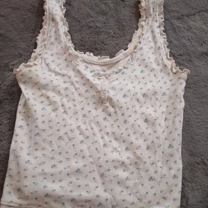 Vit blommig linnetopp från Primark - Söt vit linnetopp från Primark med små blå blommor och spetsdetaljer vid hals och ärmar. Toppen har knappar framtill och är perfekt för en avslappnad stil.