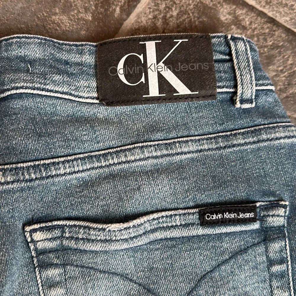 Snygga jeansshorts från Calvin Klein i en klassisk blå denim. De har en avslappnad passform med slitna detaljer och fransiga kanter för en trendig look. Shortsen har fem fickor och en knappgylf med CK-logga. Perfekta för en avslappnad stil. Står storlek 14 i men jag uppskattar dom till strl 170 för den storleken hade min son när han använde dom. . Shortsit.