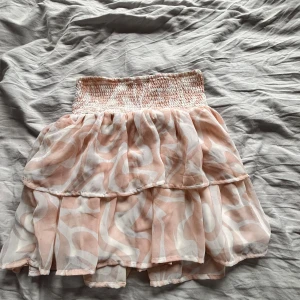 Rosa volangkjol med mönster - Söt rosa kjol med volanger och ett abstrakt vitt mönster. Den har en elastisk midja för bekväm passform och är perfekt för varma dagar. Kjolen är kort och har en luftig design.