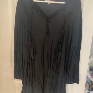 Svart långärmad klänning - Säljer en elegant svart långärmad klänning med knytband och dekorativa detaljer. Perfekt för en stilren look. Klänningen har en lös passform och är tillverkad i ett mjukt material.