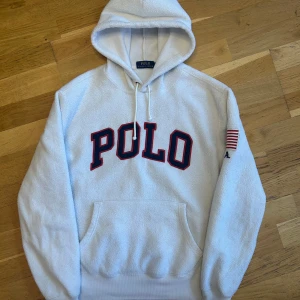 Vit hoodie från Polo Ralph Lauren - Säljer en vit hoodie från Polo Ralph Lauren med stor POLO-text i rött och blått på framsidan. Tröjan har en amerikansk flagga och texten U.S.A. på ärmen.