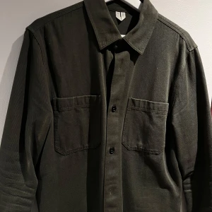 Grön overshirt från arket med fickor - Snygg grön overshirt med två stora bröstfickor och knäppning framtill. Perfekt för lager-på-lager-stil. Den har en klassisk krage och är tillverkad i ett robust material som ger en cool look. Som storlek S/M.