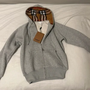Grå hoodie från Burberry - Säljer en stilren grå hoodie från Burberry med dragkedja och klassiskt rutigt mönster i luvan. Perfekt för en avslappnad och trendig look. Hoodien har långa ärmar och ribbade muddar vid ärmslut och nederkant.