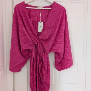 Rosa plisserad kimono från NLY Trend - Säljer en snygg rosa plisserad klänninh från NLY Trend. Den är "kvistad" framtill och vida ärmar som ger en elegant look. Perfekt för att lägga till en färgklick i garderoben! Helt ny med prislappen kvar 